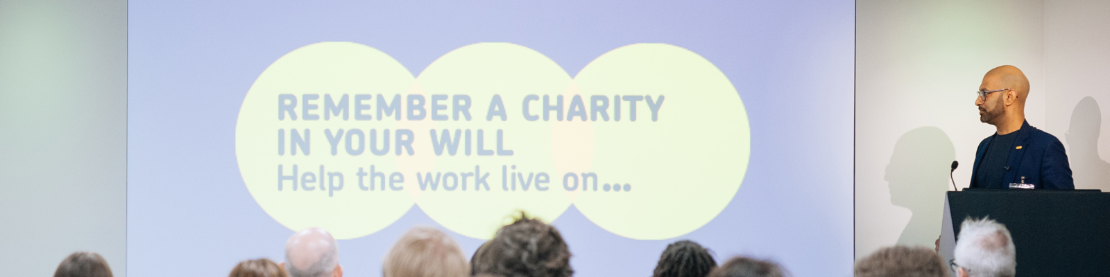 Image of a room full of delegates listening to a speaker and looking at a slide with the Remember a Charity logo on it