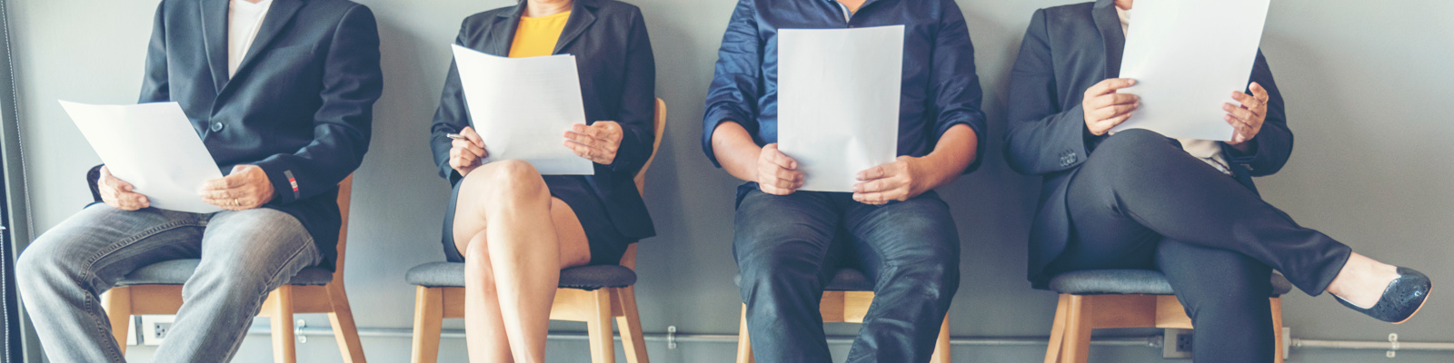 Photo of 4 candidates sitting down holding a sheet of paper preparing for a job interview. The photo cuts off the top half of their bodies showing only their legs and floor.