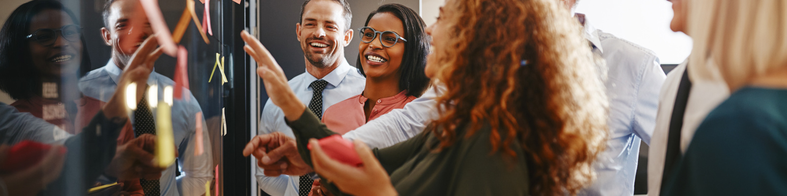 Diverse group of 6 business people laughing while brainstorming together putting sticky notes on a board.